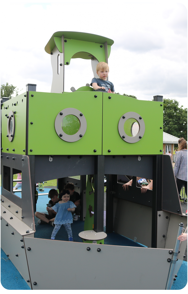 A young boy sits at the front of our lime green & grey tugboat multiplay unit. Children are also under the deck, playing together.