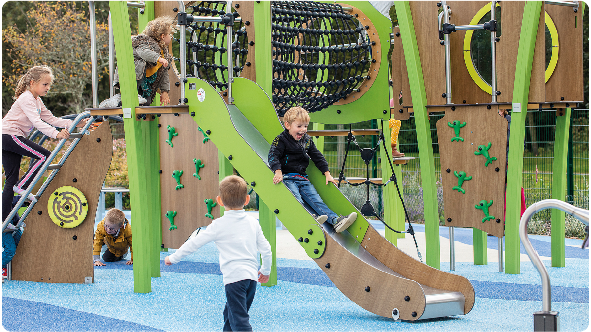 A group of young children play on a Proludic multiplay unit. A small boy in the foreground is sliding down the slide with a big grin on his face.