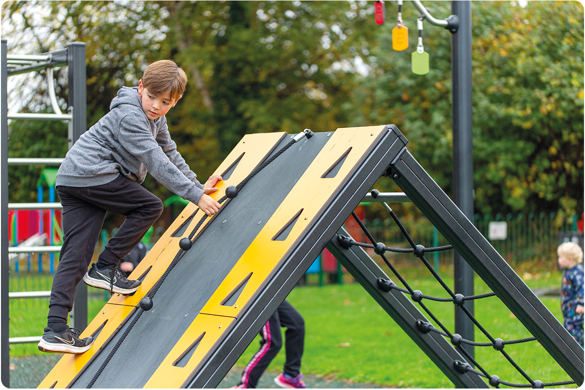 A young boy scales part of our ACTI'Ninja obstacle course equipment. The ramp has bright yellow edges with a black central board and rope. 